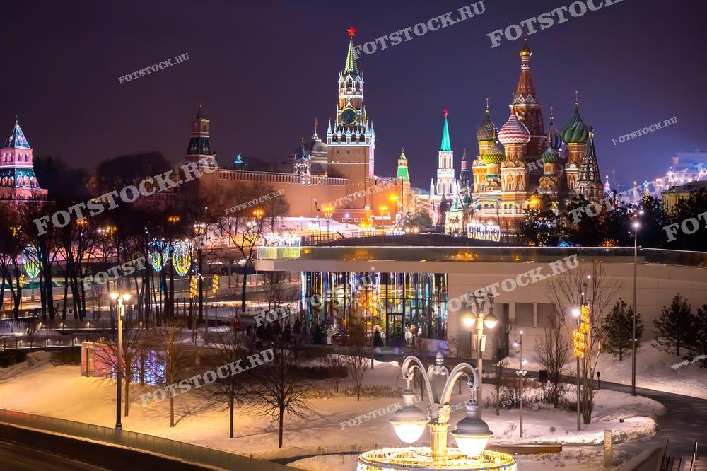 На фотографии запечатлен ночной вид на Красную площадь с видом на Собор Василия Блаженного и Спасскую башню Кремля. Эти исторические достопримечательности украшены праздничной иллюминацией, которая создает сказочную атмосферу. На переднем плане виден Парк Зарядье, также украшенный огнями. Зимняя ночь в Москве передает атмосферу праздника и новогоднего волшебства. Снег на земле и светящиеся гирлянды подчеркивают торжественность момента.